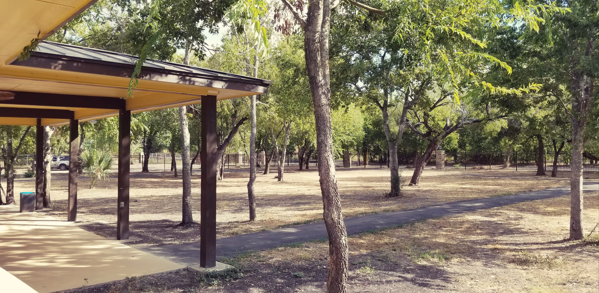 Outdoor view of a shaded patio area with brown wooden posts and a yellow ceiling, adjacent to a walking path surrounded by trees and dry grass in a fenced yard.