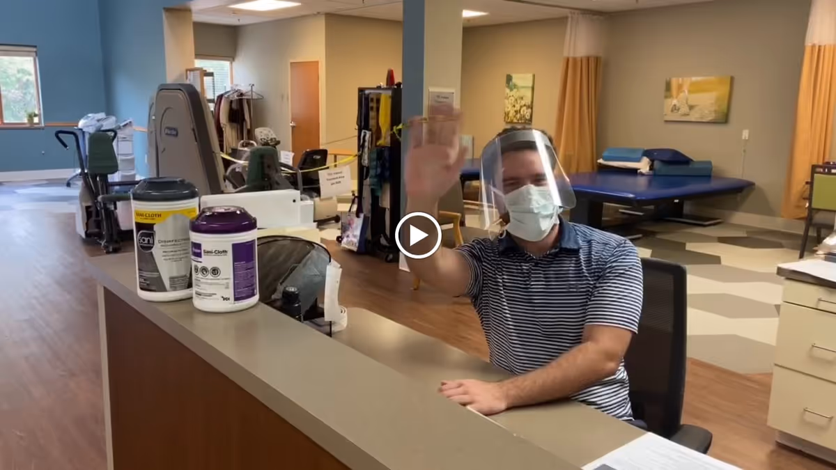 A man wearing a face mask and a clear face shield is sitting behind a reception desk in a healthcare or rehabilitation facility. He is waving and smiling. The room has exercise equipment, a treatment table, and medical supplies visible in the background.