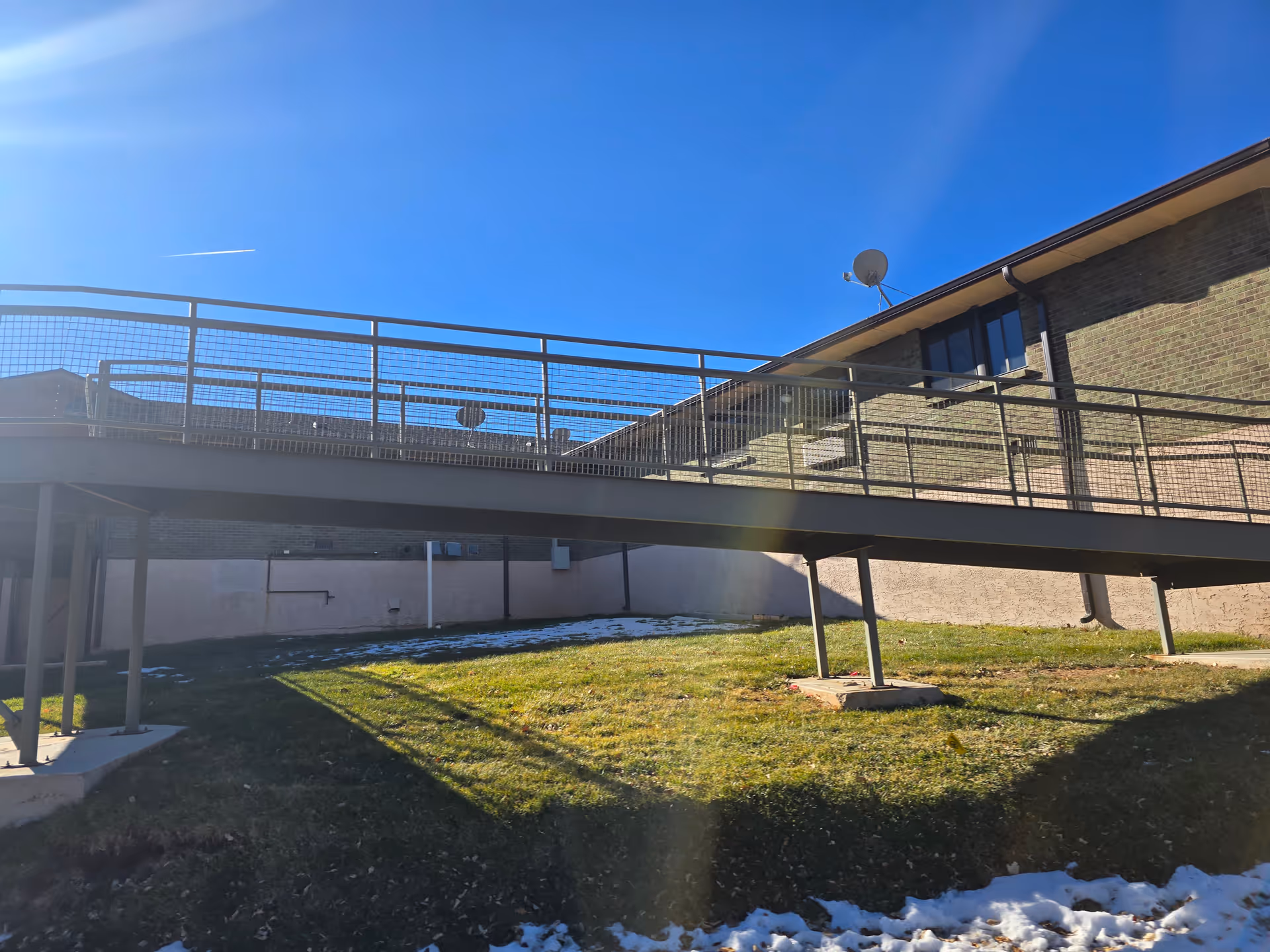 Metal elevated walkway attached to a brick building above a grassy yard with patches of snow under a clear blue sky.