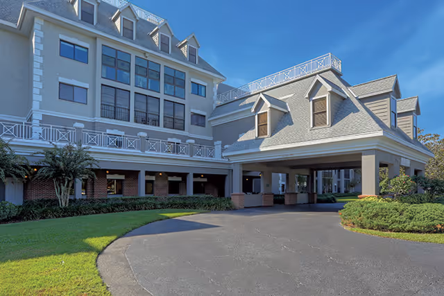 Front entrance and porte-cochère of a multi-story senior living building with dormer windows, balconies, and a circular driveway.