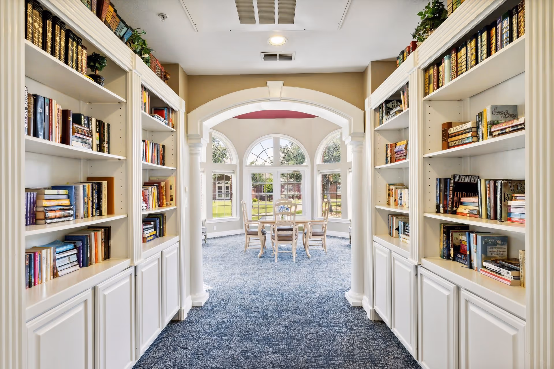 A bright and inviting interior hallway lined with white bookshelves filled with books on both sides, leading to a sunlit room with large arched windows and a small table with four chairs. The room has blue patterned carpet and beige walls with white trim.