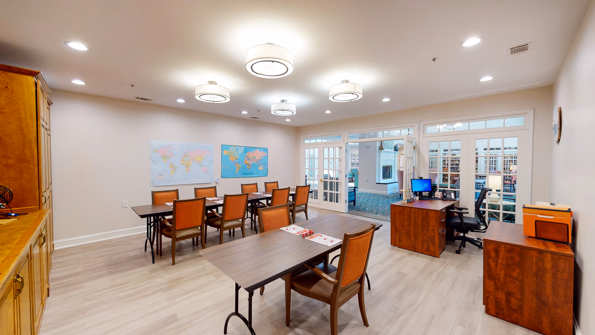 Well-lit communal activity room with multiple tables and chairs, desks with a computer, world maps on the wall, and glass French doors to an adjacent lounge.