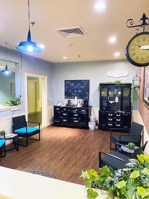 A cozy interior common area with wooden flooring, black cabinets, and wicker chairs with blue cushions. There are hanging pendant lights, a wall clock with Roman numerals, and a chalkboard sign that says 'Coffee & Tea'. Green plants are placed around the room, adding a touch of nature.