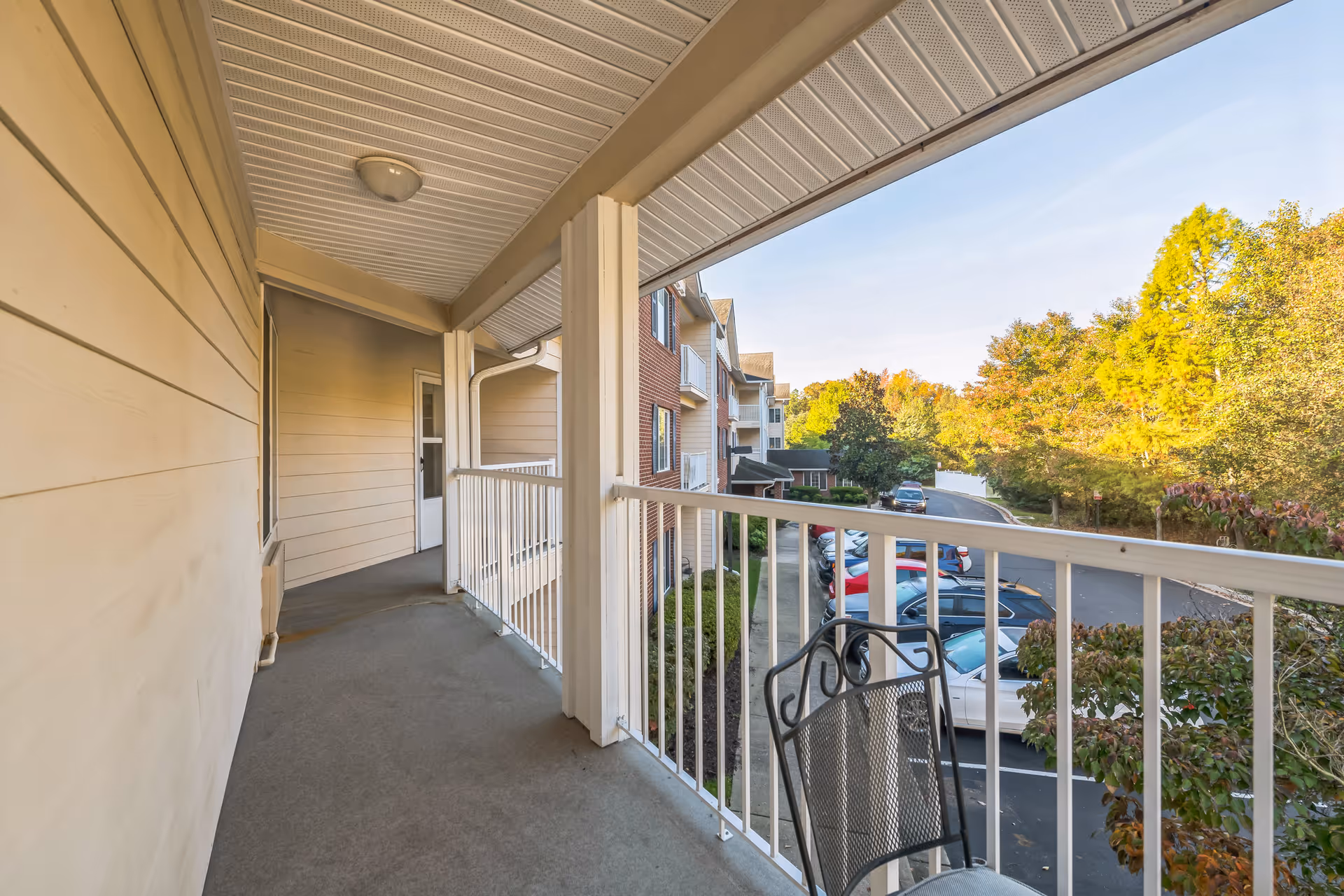 View from a covered balcony with white railing overlooking a parking lot and trees with autumn foliage. The balcony has a metal chair and is part of a multi-story building with beige siding and brick accents.