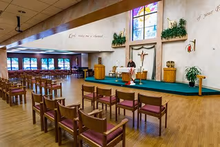 Interior view of a chapel or worship area with rows of wooden chairs with red cushions, a raised platform with a lectern, a crucifix on the wall, stained glass windows, and plants decorating the space. The walls have inspirational text written on them.
