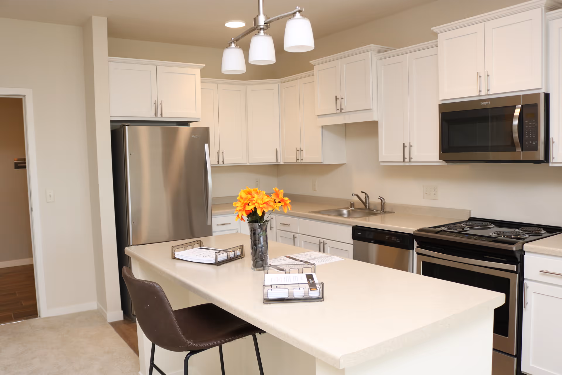 Modern kitchen with white cabinets, stainless steel refrigerator, microwave, dishwasher, and stove. A kitchen island with a brown chair and a vase of orange flowers is in the center.