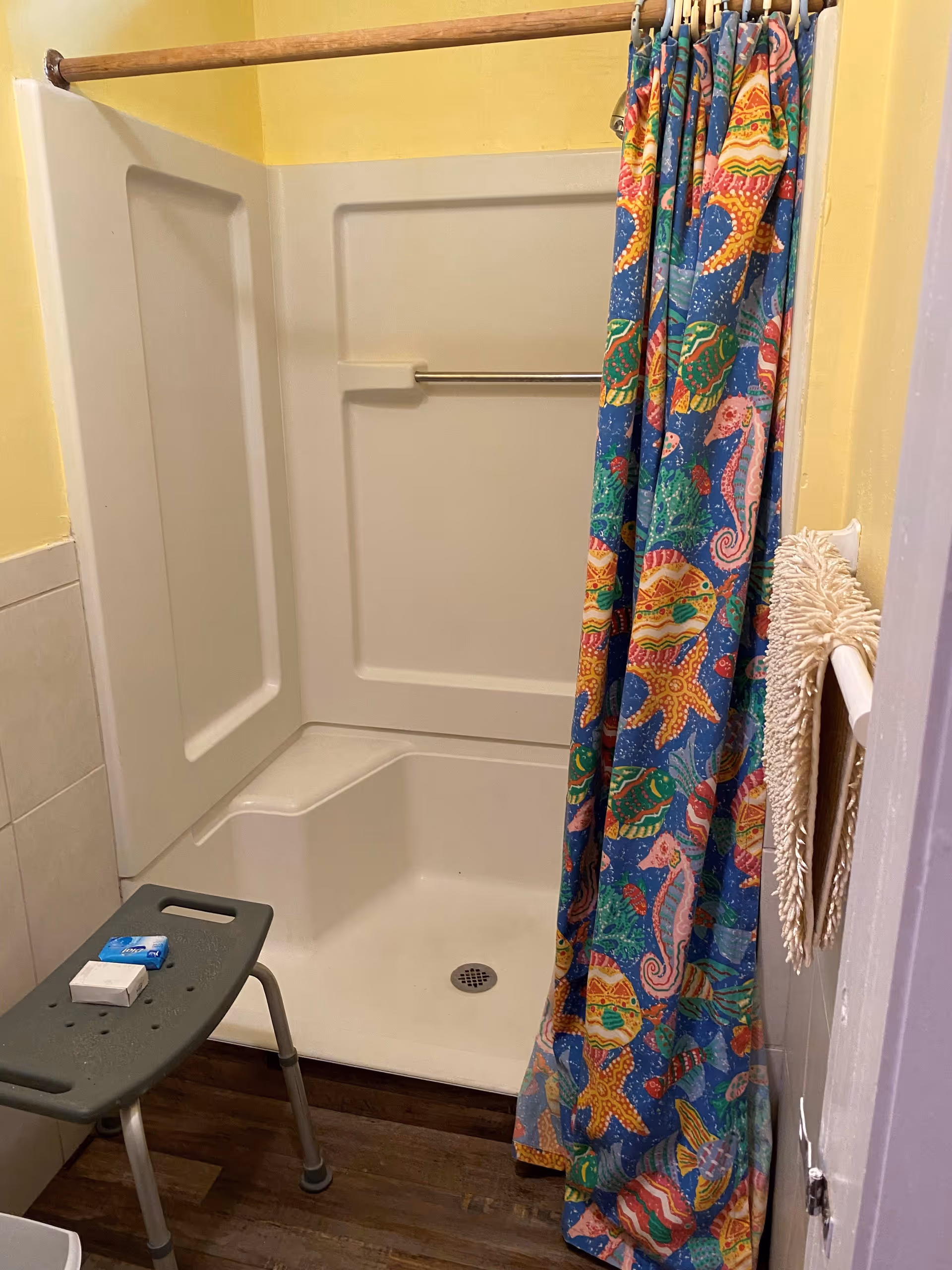 A small shower area with a white plastic shower stall and a colorful shower curtain featuring sea creatures like starfish and seahorses. There is a gray shower bench with two bars of soap on it. The walls are painted yellow and there is a towel hanging on a rack on the right side.