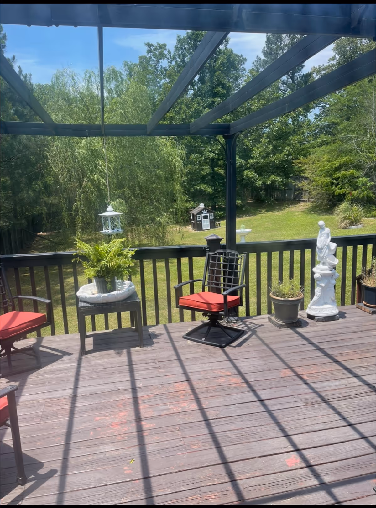 Outdoor patio area with wooden deck flooring, black metal railing, and a pergola overhead casting shadows. There are two black metal chairs with red cushions, a small table with a potted fern, a white hanging lantern, several potted plants, and a white statue. In the background, there is a grassy yard with trees and a small playhouse.
