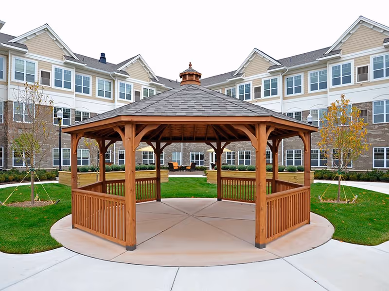 A wooden gazebo with a shingled roof situated in the center of a circular concrete area surrounded by green grass and young trees, with a multi-story senior living building in the background.