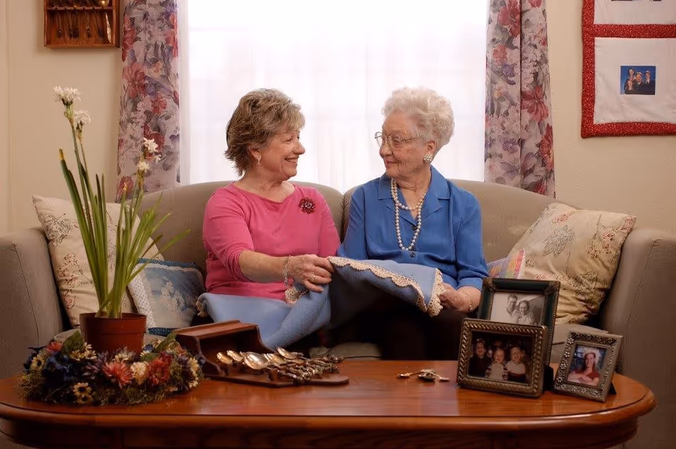 Two elderly women sitting on a beige couch in a cozy living room, smiling at each other while holding a blue blanket. The room has floral curtains, framed family photos on a wooden coffee table, and decorative pillows on the couch.