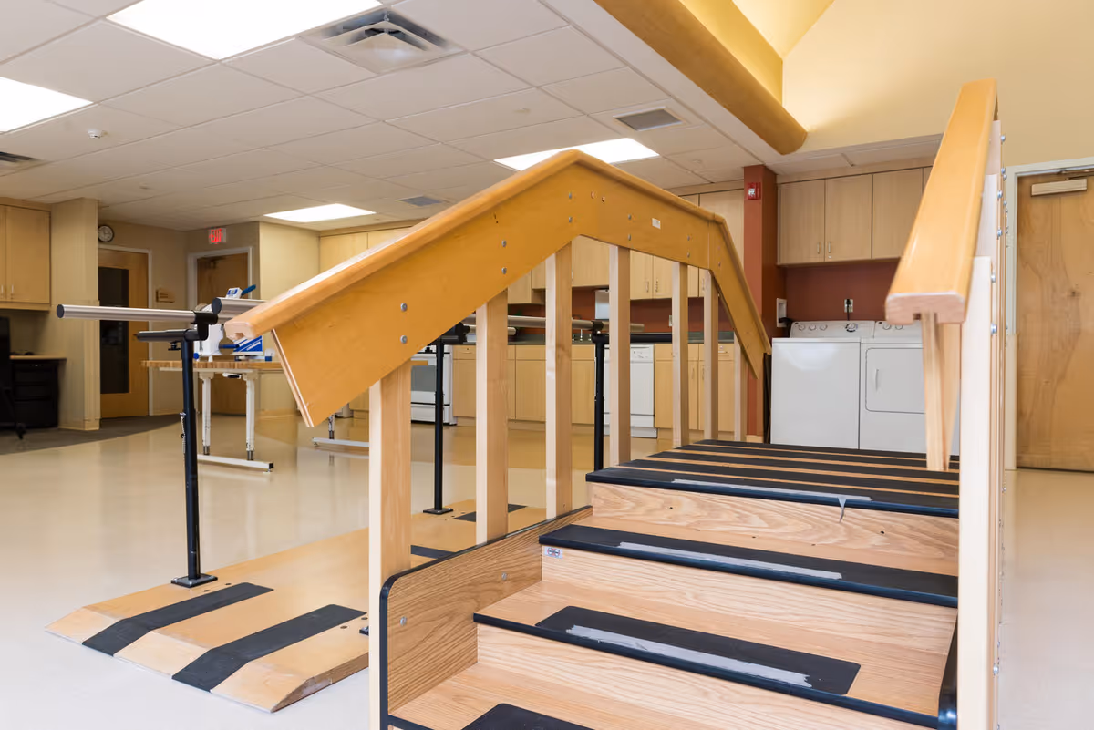 Indoor rehabilitation room with wooden stairs and ramp equipped with handrails for physical therapy. The background shows a kitchen area with cabinets, a stove, a dishwasher, and a washer and dryer unit.