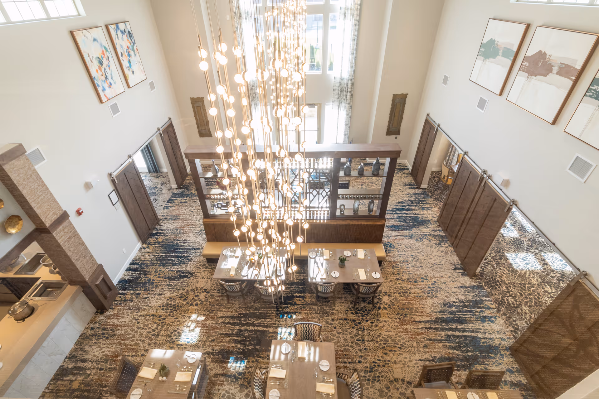 High-ceiling communal dining area with multiple set tables, a large cascading chandelier, decorative shelving and patterned carpet.