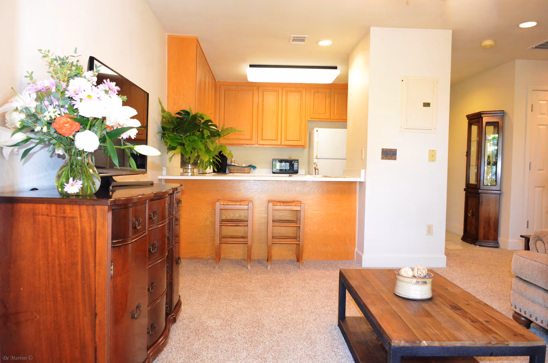Interior view of a living room and kitchen area in a retirement living community. The living room features a wooden dresser with a vase of flowers and a flat-screen TV on top, a wooden coffee table with decorative items, and a beige upholstered chair. The kitchen has wooden cabinets, a white refrigerator, a microwave, and a counter with two wooden stools. There is a tall wooden display cabinet in the hallway area.