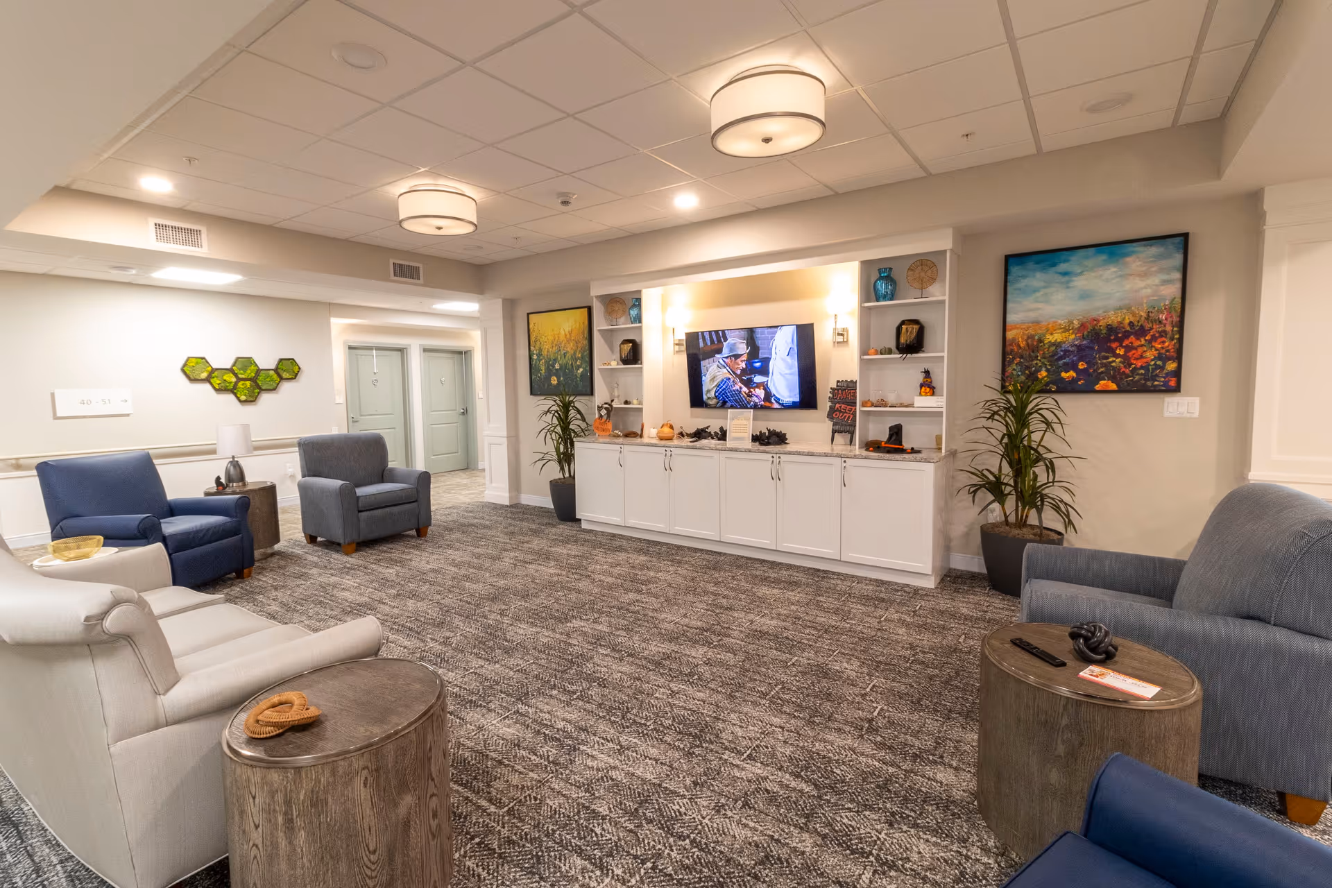 Bright communal living room/lounge with upholstered chairs, a TV set into white shelving, side tables, and colorful artwork.