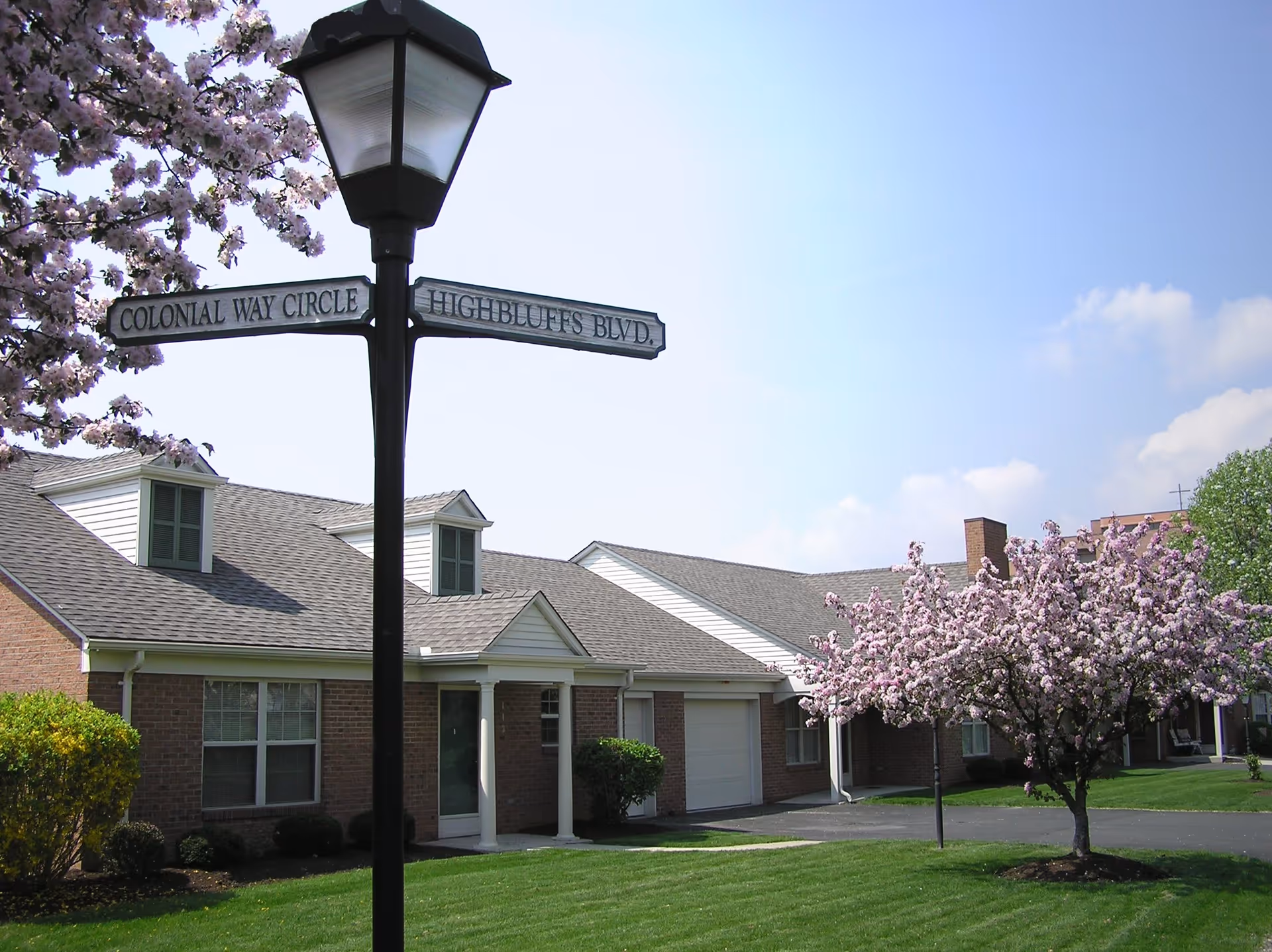 A street lamp post with two street signs reading 'COLONIAL WAY CIRCLE' and 'HIGHBLUFFS BLVD.' in front of a single-story brick building with white trim and dormer windows. There is a blooming tree with pink flowers and well-maintained green grass under a clear blue sky.