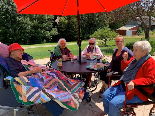 Four elderly women and one younger woman sitting around a round outdoor table under a large red umbrella. The elderly women are seated in wheelchairs and are covered with blankets. They are enjoying drinks and snacks on a sunny day with green trees and grass in the background.