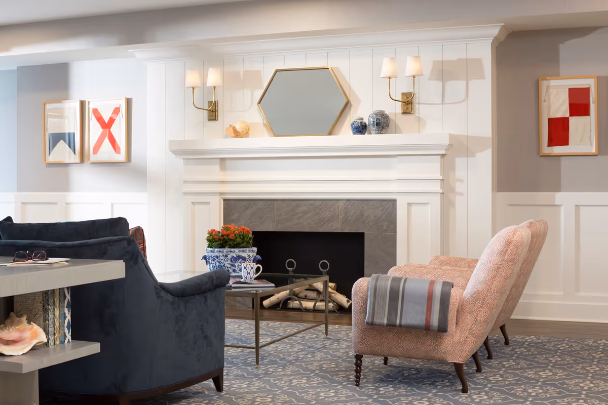 A cozy living room area featuring a white fireplace with a hexagonal mirror and decorative vases on the mantel. Two wall sconces with lampshades flank the mirror. In front of the fireplace, there is a glass coffee table with a blue and white floral planter holding red flowers, a cup, and some books. To the left, a dark blue armchair is partially visible, and to the right, a patterned pink armchair with a folded gray and red blanket draped over the arm. The room has framed abstract art on the walls and a patterned blue carpet.