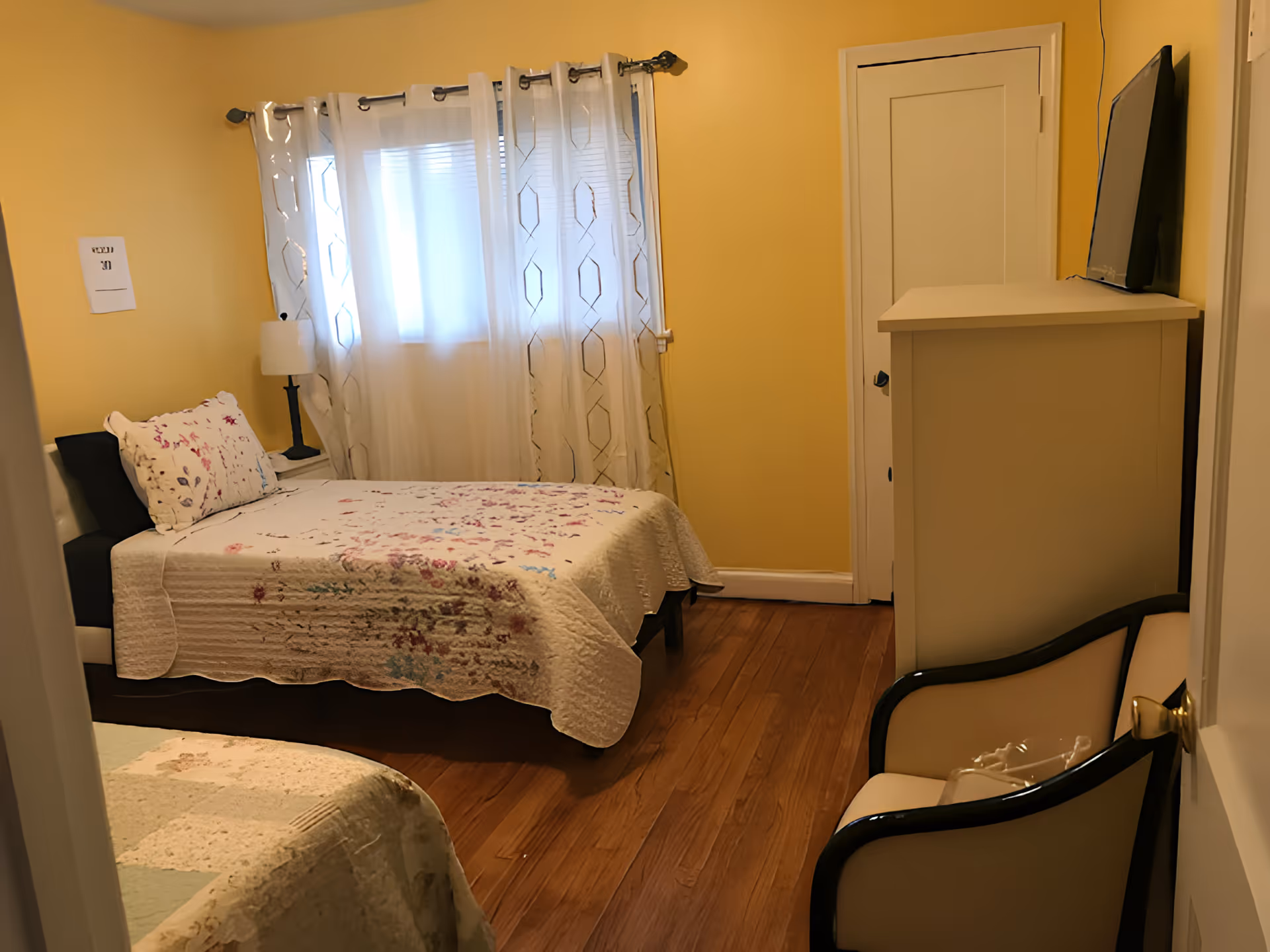 A small bedroom with yellow walls and wooden floor. The room contains two beds with floral bedspreads, a white dresser with a TV on top, a white armchair with black armrests, a window with sheer white curtains, and a small bedside table with a lamp.