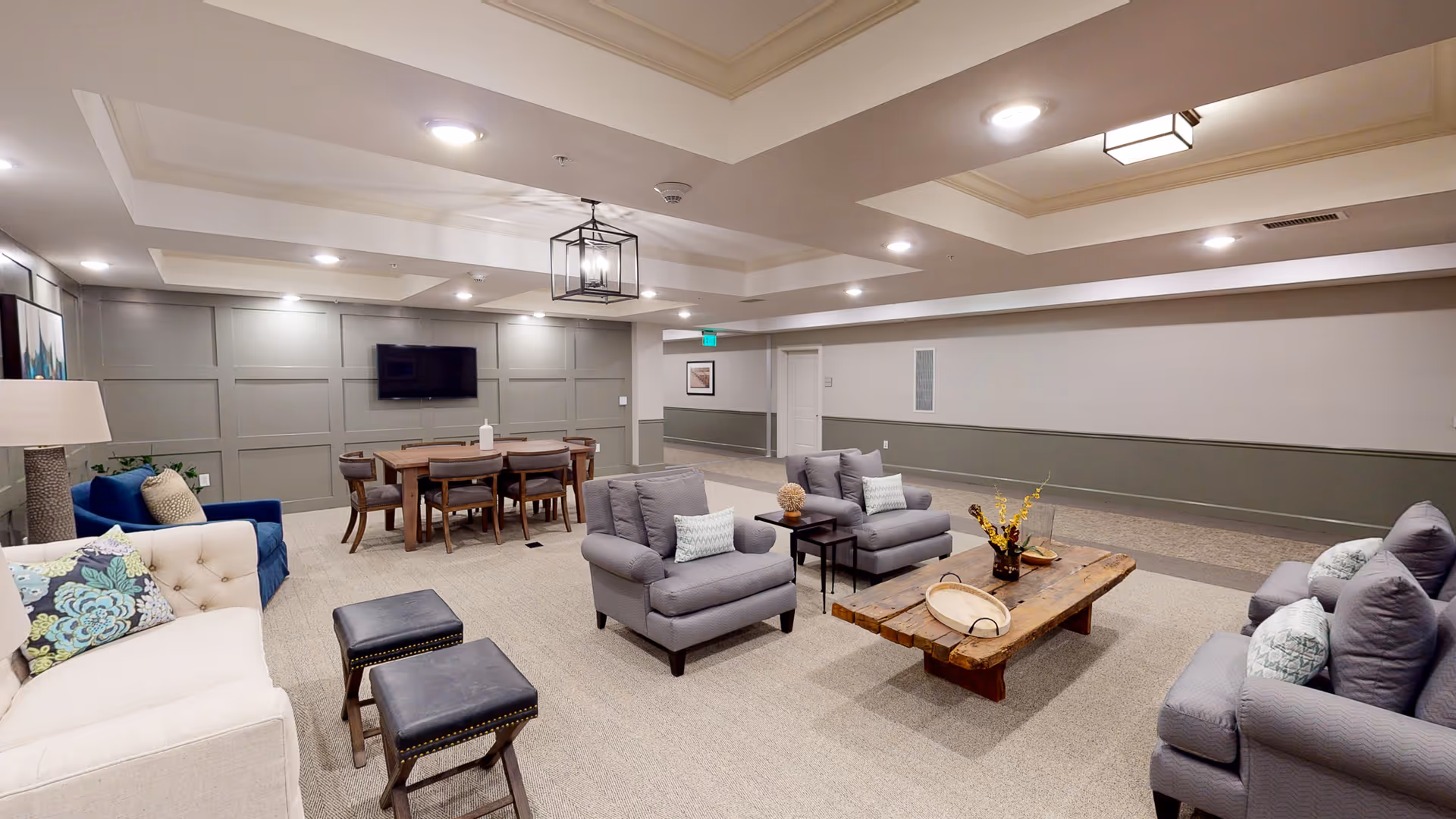 A spacious lobby area featuring comfortable seating, a wooden coffee table, and a dining table. The walls are adorned with decorative paneling and a television is mounted on one wall.