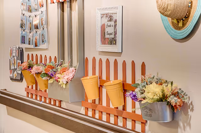 Hallway wall decorated with small red picket-fence shelves holding yellow pots of artificial flowers, framed art and a hanging hat above a handrail.