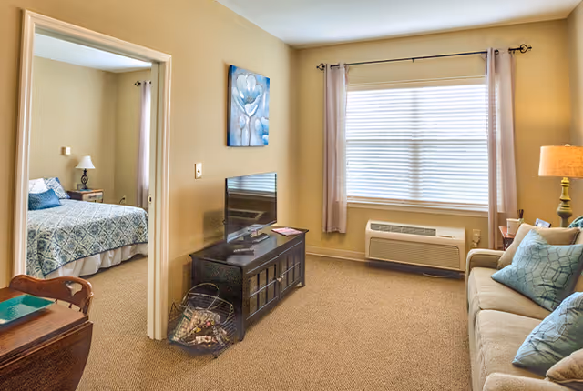 A cozy living room with a beige sofa adorned with blue and beige pillows, a floor lamp, and a window with closed blinds and light curtains. A black TV stand with a flat-screen TV is positioned against the wall, and a doorway leads to a bedroom with a bed covered in a blue patterned bedspread and a bedside table with a lamp.