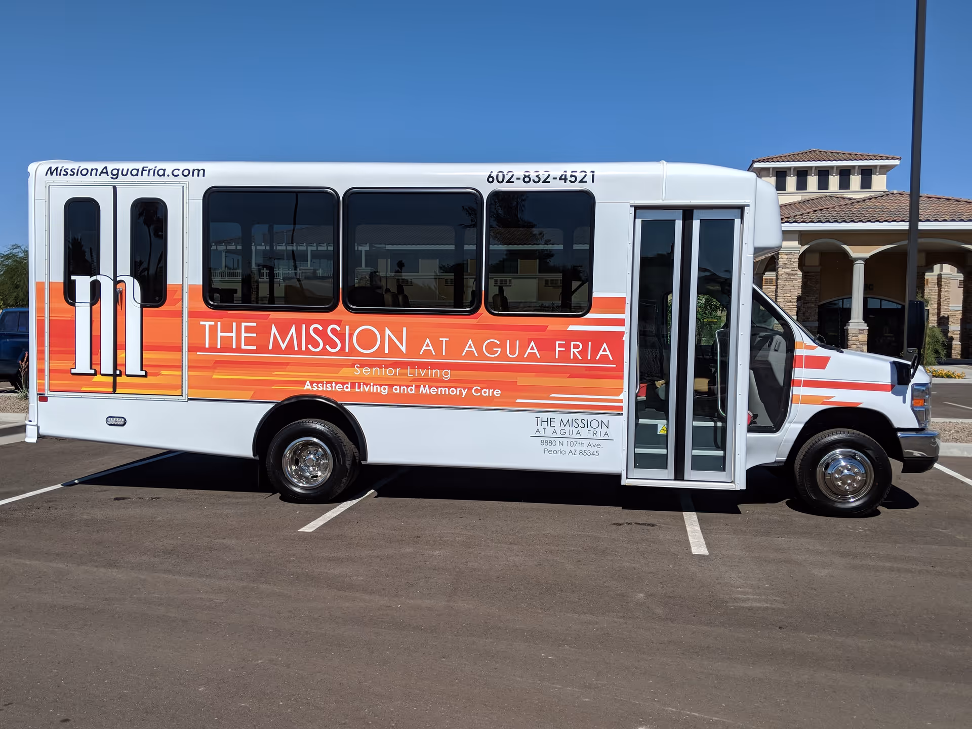 A white shuttle bus with orange and red stripes parked in a lot. The bus has text on the side that reads 'THE MISSION AT AGUA FRIA Senior Living Assisted Living and Memory Care' along with the website MissionAguaFria.com and phone number 602-832-4521. A building with a tiled roof is visible in the background under a clear blue sky.