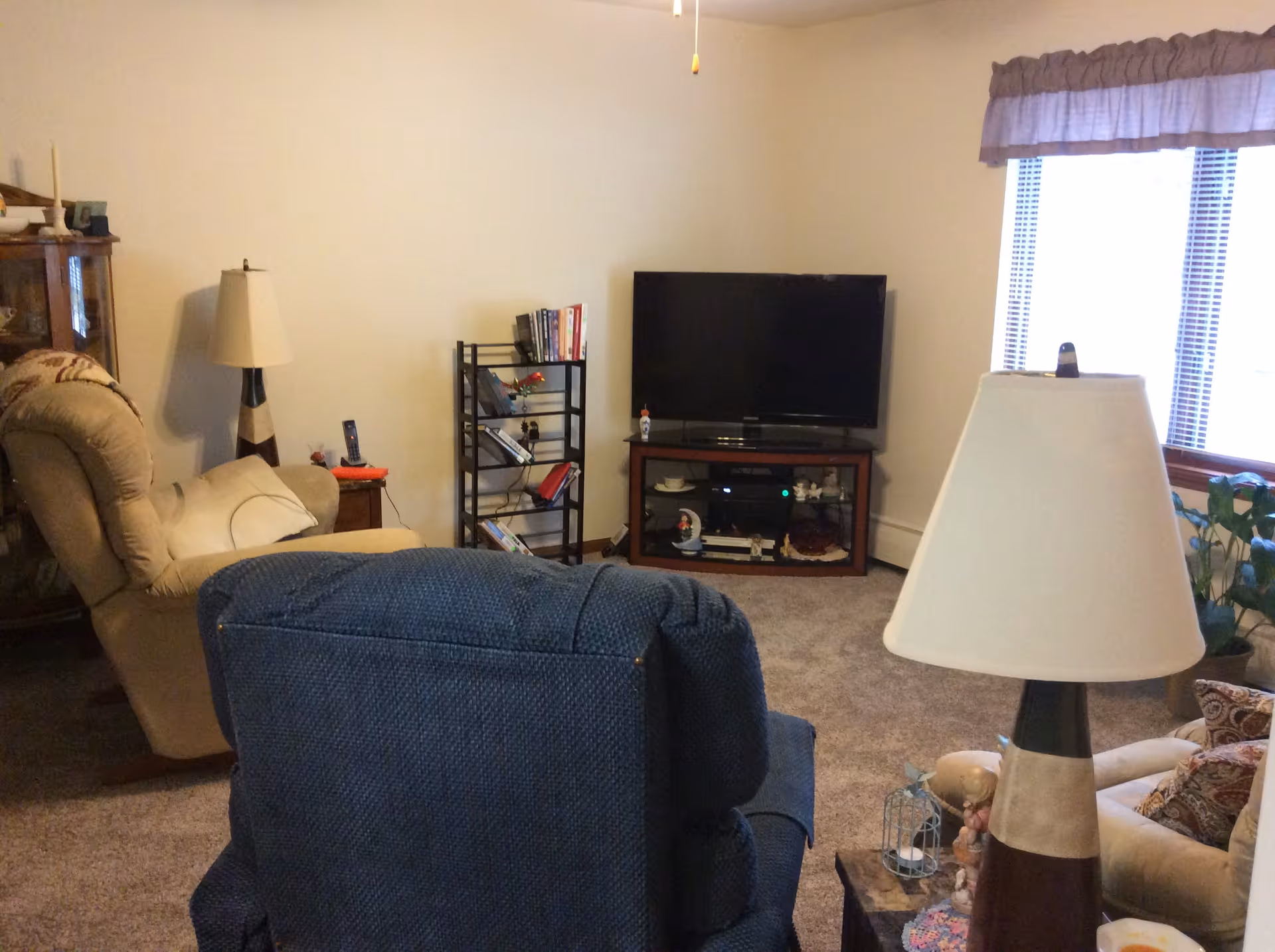 A cozy living room with a blue recliner chair in the foreground, a beige recliner chair to the left, a wooden TV stand with a flat-screen TV, a small black bookshelf with books, a floor lamp, and a window with blinds and a valance letting in natural light.