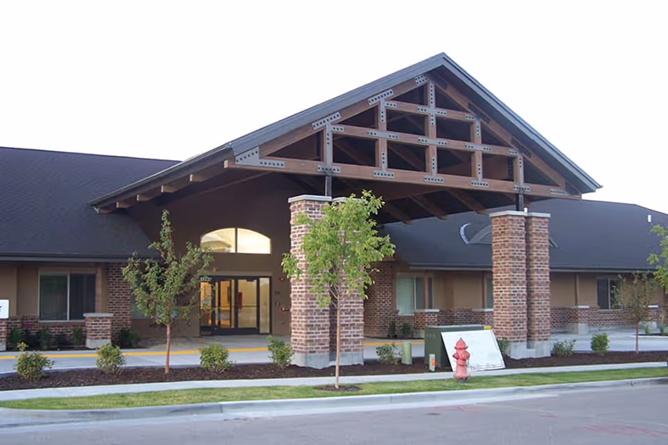 Front entrance of a single-story brick building with a large wooden portico, glass doors, and small trees and shrubs.