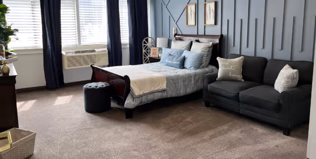 A well-lit bedroom with a large window covered by white blinds and dark curtains. The room features a wooden bed with light blue bedding and multiple pillows, a dark gray loveseat with decorative pillows, a small round ottoman, and framed artwork on a textured blue accent wall.
