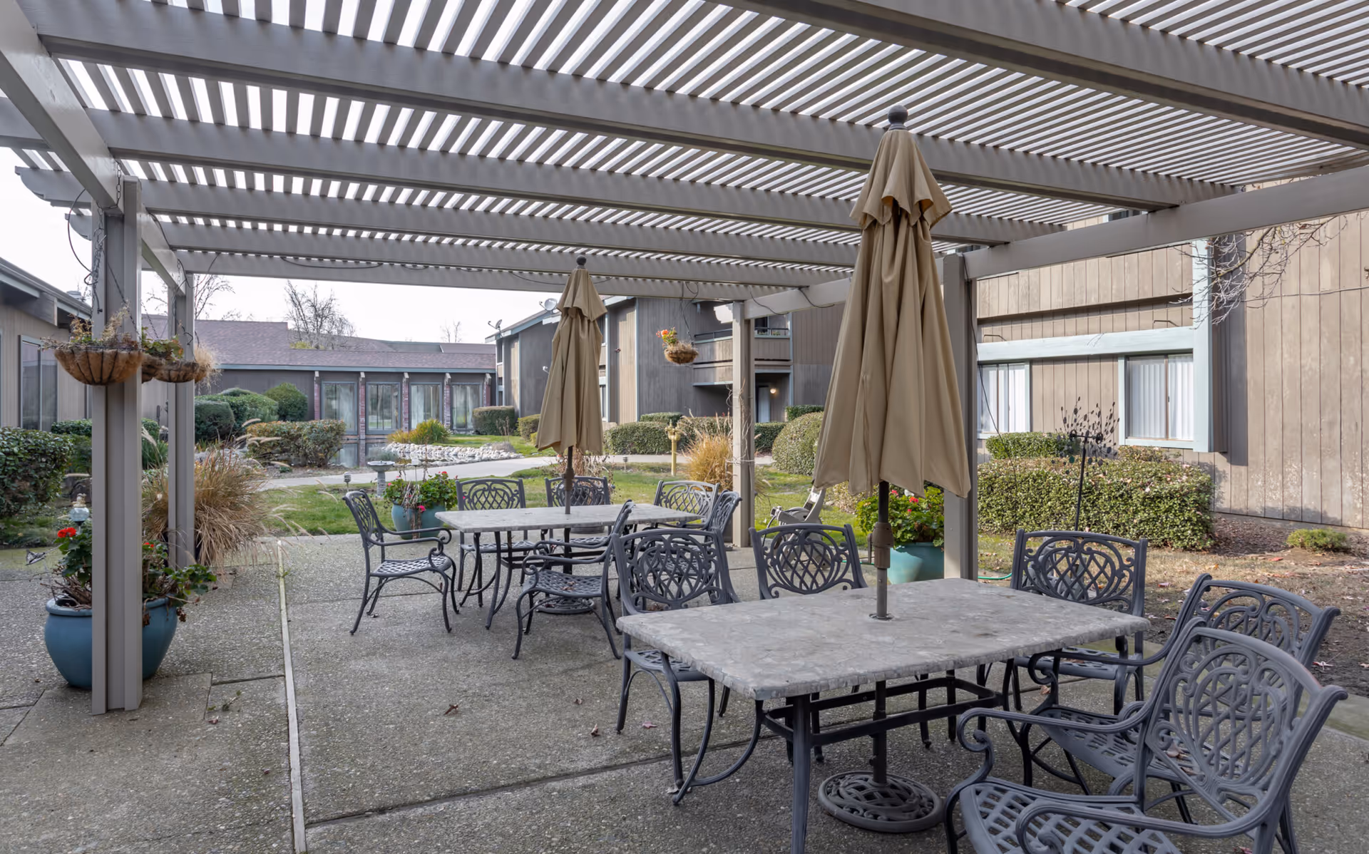 Outdoor patio area with metal tables and chairs under a pergola. Two tables have closed beige umbrellas in the center. Surrounding the patio are potted plants and bushes, with a building visible in the background.