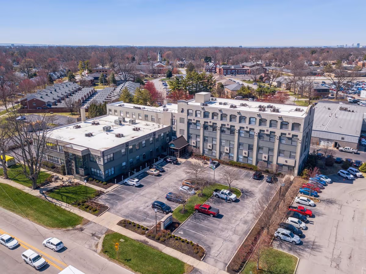 Aerial view of a multi-story building with a parking lot in front, surrounded by trees and residential houses in the background under a clear sky.