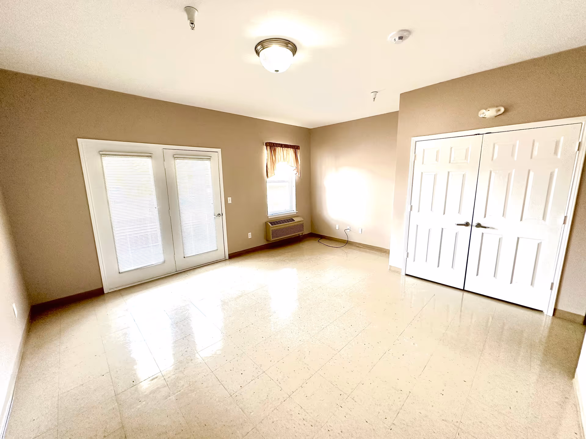 Empty room with beige walls and tiled floor, featuring a set of white double doors on the right, a window with a brown valance and an air conditioning unit below it, and white double French doors with blinds on the left side.