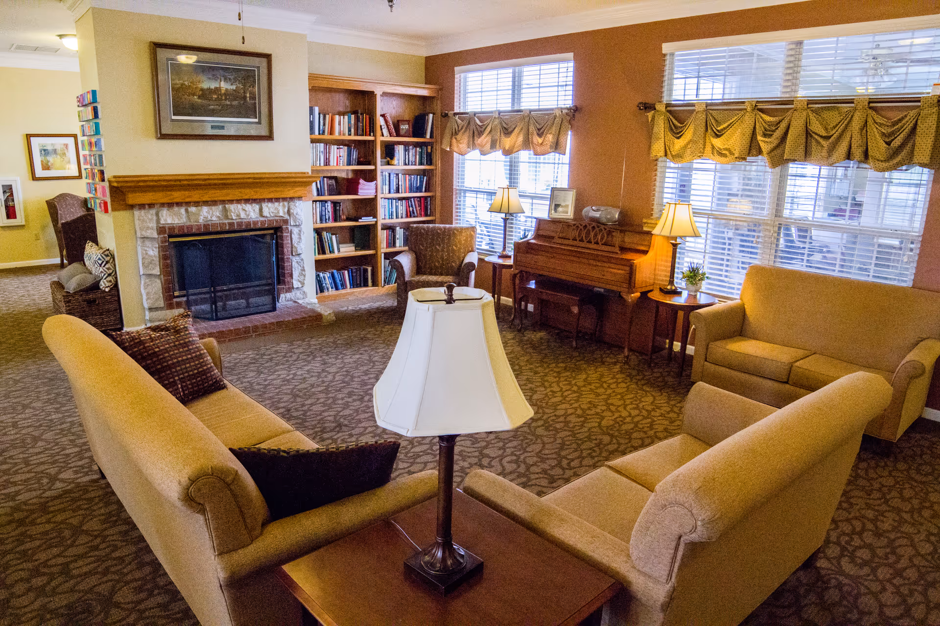 Cozy senior living lounge with sofas and armchairs arranged around a central lamp, a fireplace, bookshelf, and a piano beside large windows.