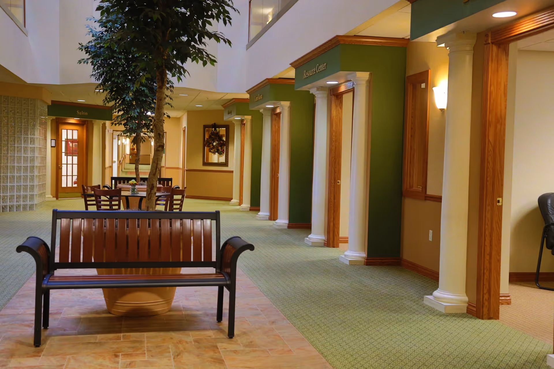 Interior hallway of Maple Ridge Retirement Community featuring a wooden bench in the foreground, a potted tree behind it, and a round table with chairs further back. The hallway has green carpet, cream walls with wooden trim, white columns, and signs above doors indicating rooms such as the Resource Center and Salon.