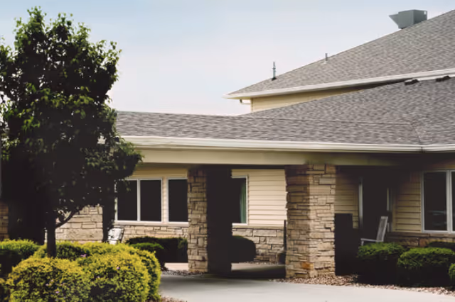 Exterior view of a senior living facility building with stone and siding walls, a covered entrance supported by stone pillars, and surrounding bushes and a tree.