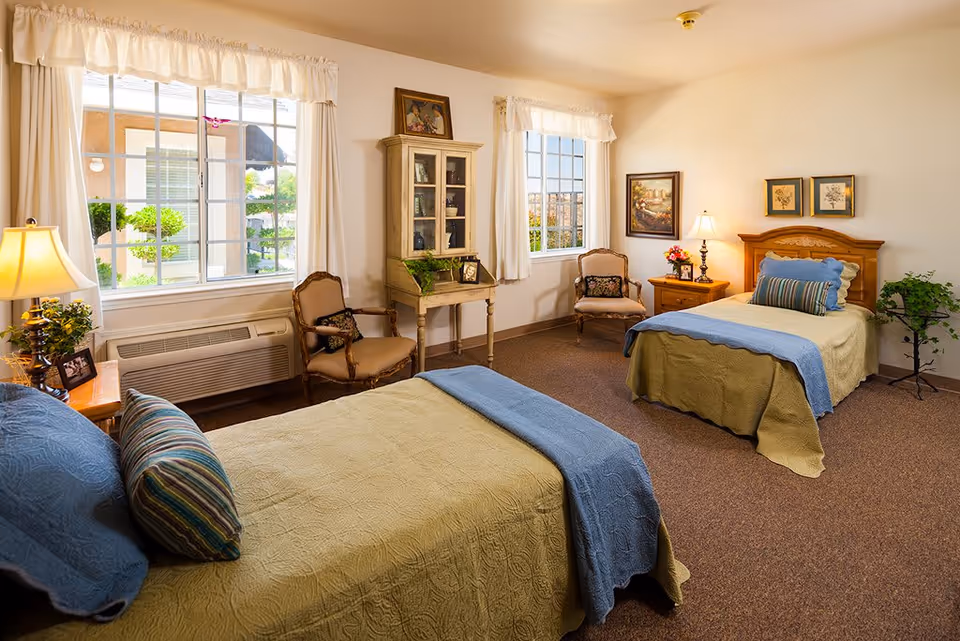 A cozy bedroom with two twin beds, each with a green bedspread and blue blanket. The room has two windows with white curtains, two wooden chairs with cushions, a wooden cabinet with glass doors, a nightstand with a lamp and flowers, framed artwork on the walls, and a small plant in the corner.