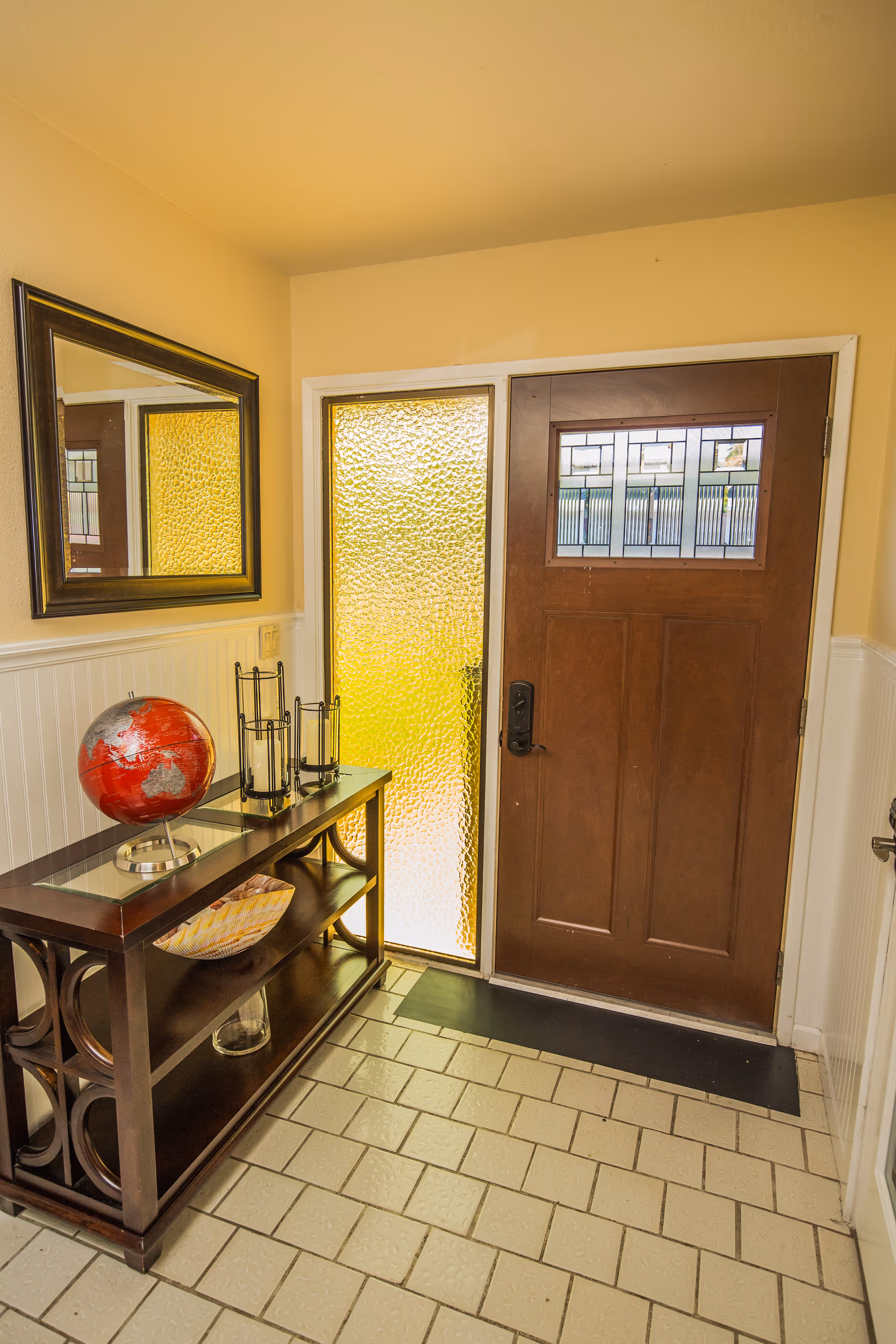 Entryway with a wooden front door featuring a decorative glass panel, a side window with textured glass, a dark wooden console table with a red globe, candle holders, and a basket, a large rectangular mirror on the wall, and tiled flooring.