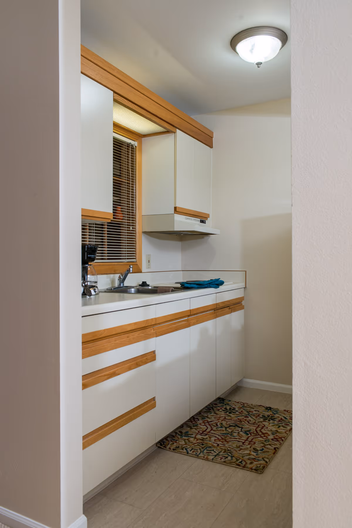A small kitchen area with white cabinets featuring wooden handles, a countertop with a sink, a coffee maker, and a window with blinds. There is a patterned rug on the floor and a ceiling light fixture above.