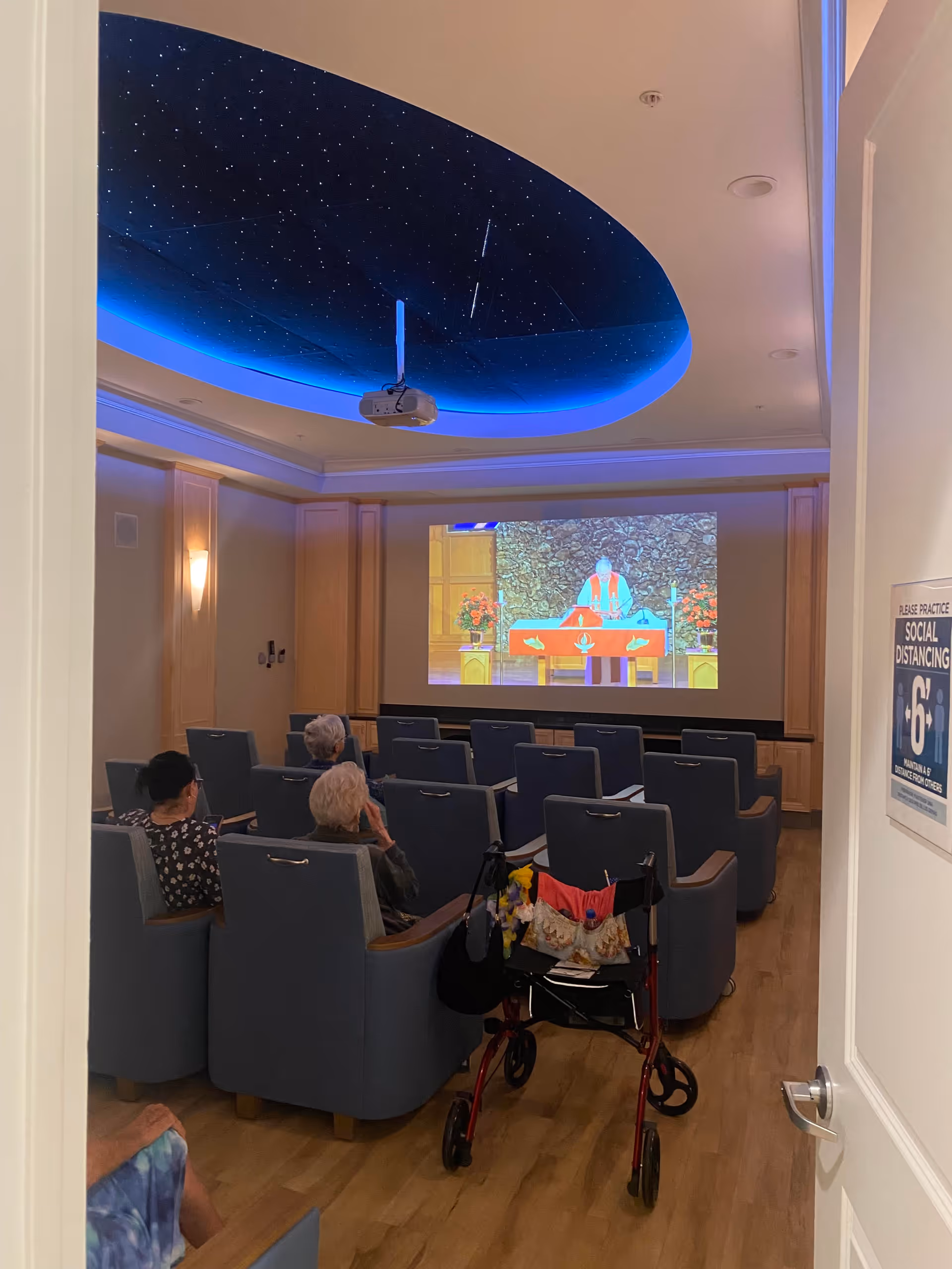 A small group of elderly people seated in a room with blue chairs watching a religious service projected on a screen. The ceiling has a starry night design with blue lighting. A red walker is parked near the chairs. A sign on the door reminds to practice social distancing of 6 feet.