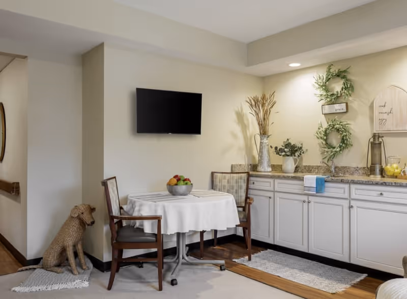 A cozy corner in a senior living facility with a small round table covered with a white tablecloth, two wooden chairs, a wall-mounted flat screen TV, and a countertop with decorative plants and wreaths on the wall above. A woven dog statue sits on a rug in the corner.