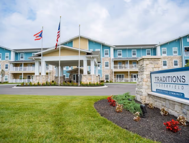 Front exterior of the Traditions of Deerfield senior living community showing a covered entrance, three flagpoles, landscaped lawn, and a stone sign.