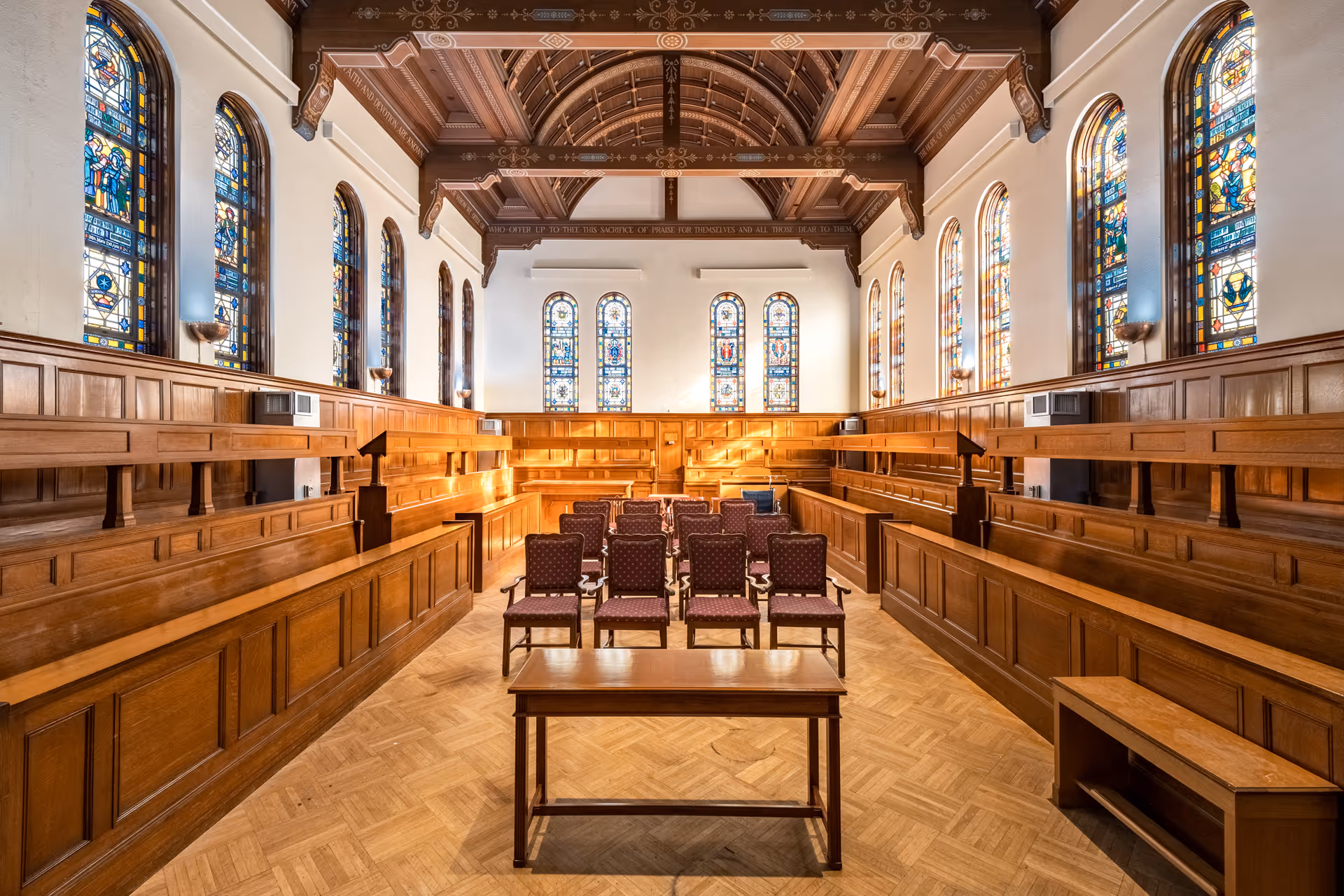 Interior of a chapel or assembly room with wooden pews arranged in tiers along the sides and back, several upholstered chairs in the center, stained glass windows on the walls, and a decorative wooden ceiling.