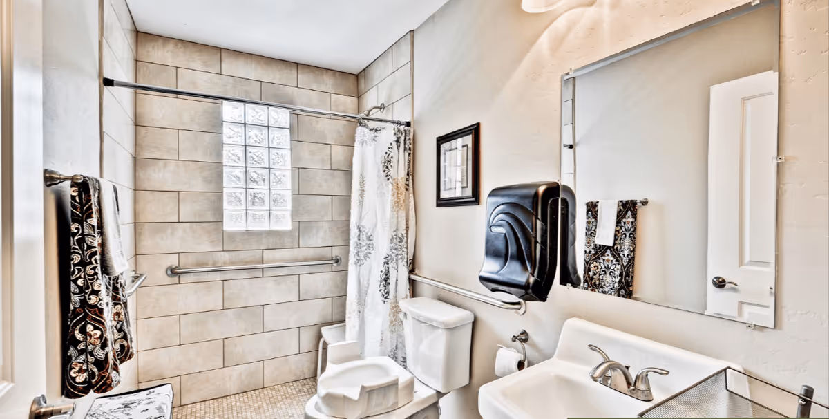 A clean and accessible bathroom featuring a tiled walk-in shower with a glass block window, a white toilet with a raised seat, a white sink with a silver faucet, a large mirror, a black wall-mounted soap dispenser, and decorative towels hanging on towel bars.