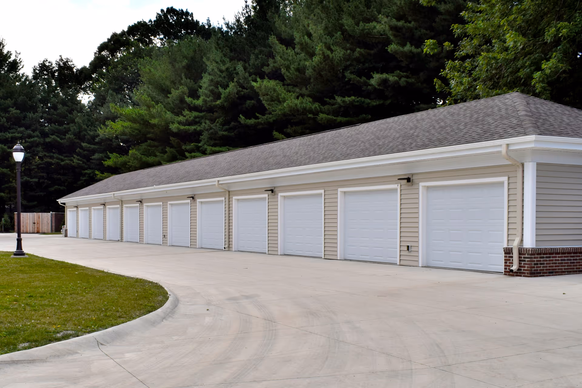 A row of closed white garage doors on a beige building with a gray shingled roof, surrounded by trees and a concrete driveway curving in front of the garages.