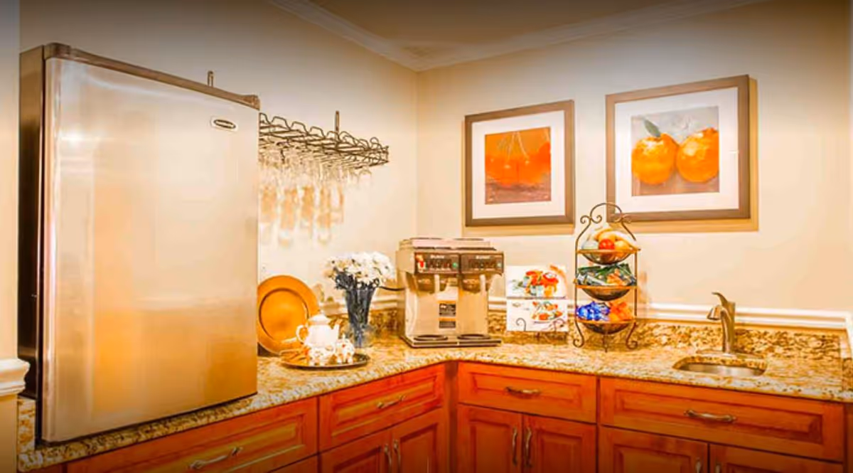 Small kitchenette or coffee bar with a stainless refrigerator, coffee machine, sink, fruit stand, and framed fruit artwork above wooden cabinets.