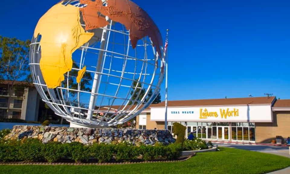 Large metal globe sculpture on a landscaped rock bed in front of a Leisure World building entrance under a clear blue sky.