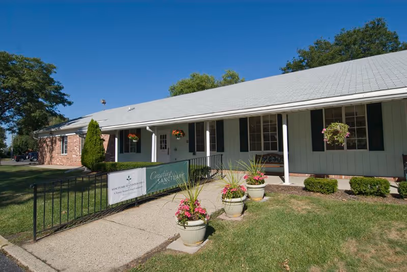 Exterior view of a single-story senior living facility building with a light gray roof, white siding, and green shutters. There is a concrete walkway leading to the entrance, lined with potted plants with pink flowers. Hanging flower baskets are visible near the entrance, and a green banner with white text is attached to a black metal railing along the walkway. The sky is clear and blue, and there are trees and grass surrounding the building.