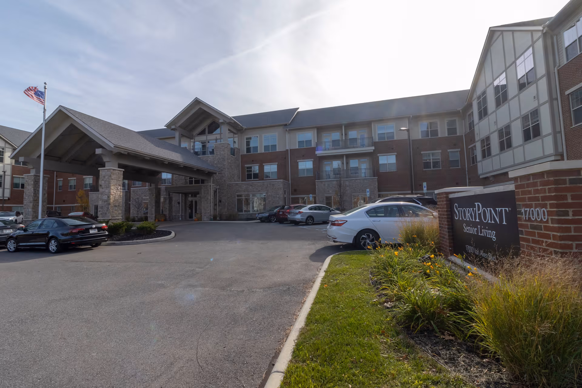Exterior view of StoryPoint Senior Living facility in Shaker Heights, showing a large three-story building with a covered entrance, several parked cars, an American flag on a flagpole, and a landscaped area with flowers and grass near the facility sign.