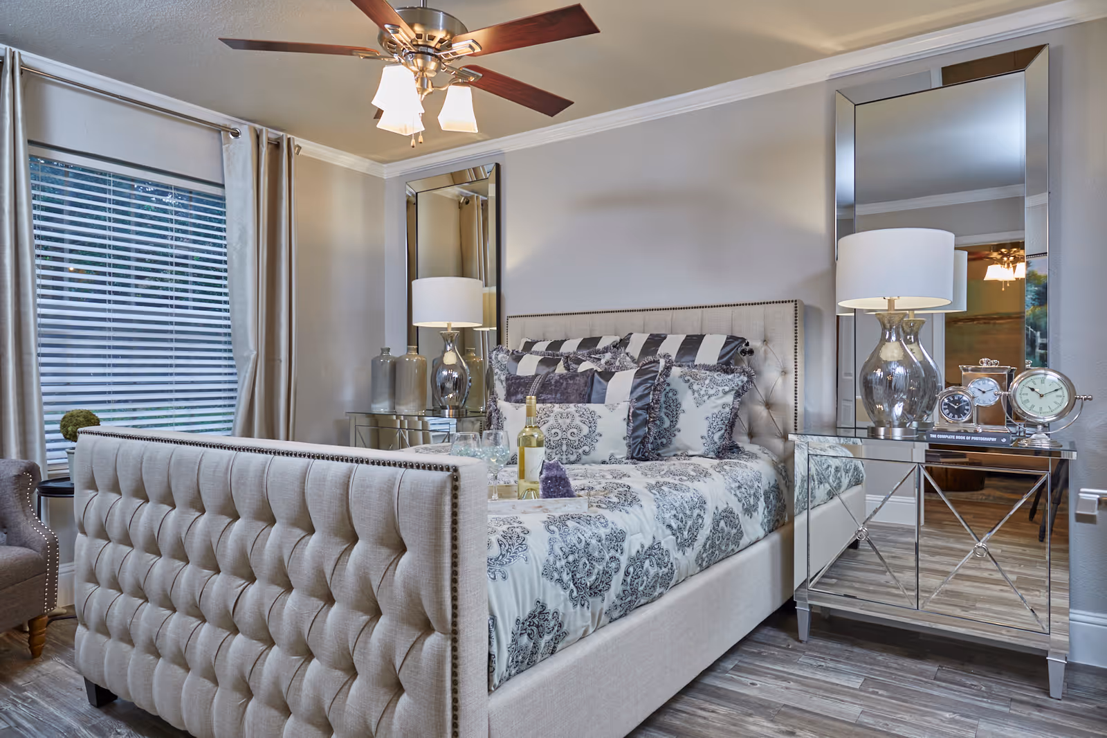 A well-decorated bedroom featuring a tufted beige upholstered bed with patterned bedding and multiple pillows. On the right side, there is a mirrored nightstand with a silver lamp, three clocks, and a book. Two tall mirrors are mounted on the wall behind the nightstand. A window with blinds and beige curtains is on the left side, next to a gray armchair with nailhead trim. The room has wood flooring and a ceiling fan with lights.
