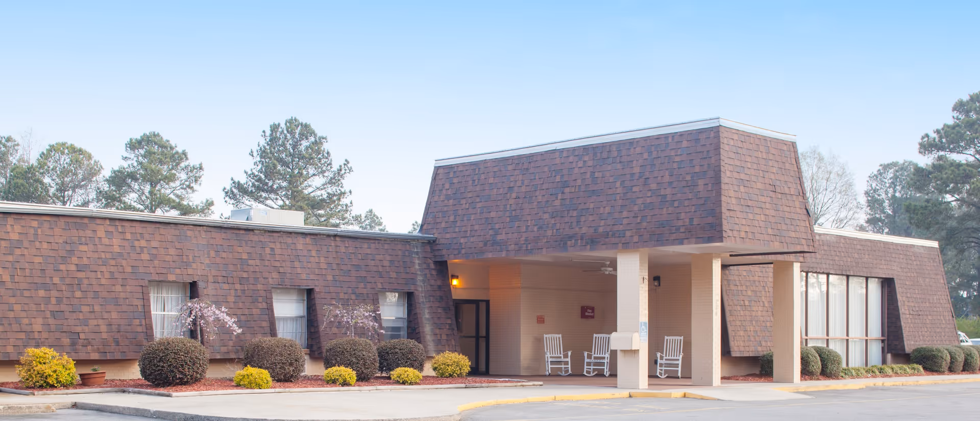 Exterior view of Mount Olive Center, a single-story building with brown shingle siding and a covered entrance with white rocking chairs. There are small bushes and trees around the building and a clear sky above.